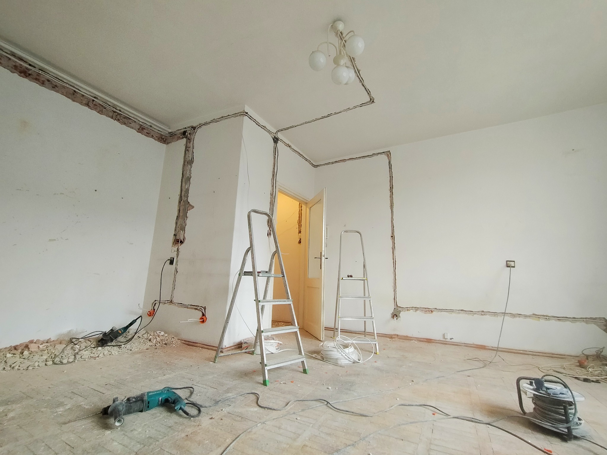 Remont instalacji elektrycznej w pokoju: bruzdy w ścianach, kable, puszki elektryczne, wiertarka udarowa na podłodze, dwie drabiny aluminiowe.