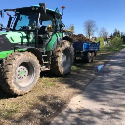 Zielony ciągnik Deutz-Fahr z przyczepą wypełnioną ziemią, stojący na poboczu asfaltowej drogi w słoneczny dzień. Widoczne błotniste opony i świeżo wykopana ziemia w przyczepie.