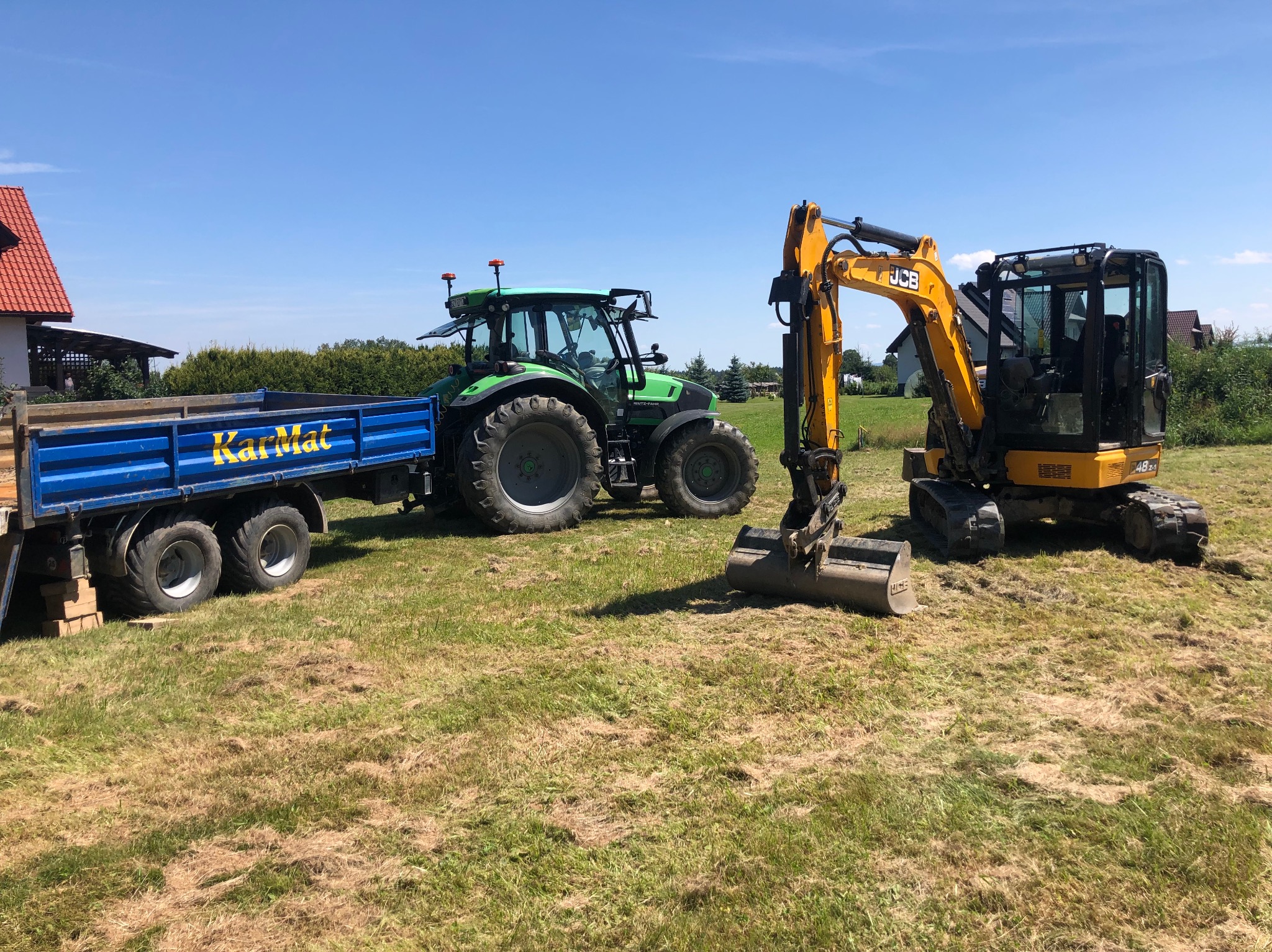 Żółta minikoparka JCB i zielony traktor Deutz-Fahr z niebieską przyczepą Karmat na trawiastym terenie w słoneczny dzień.