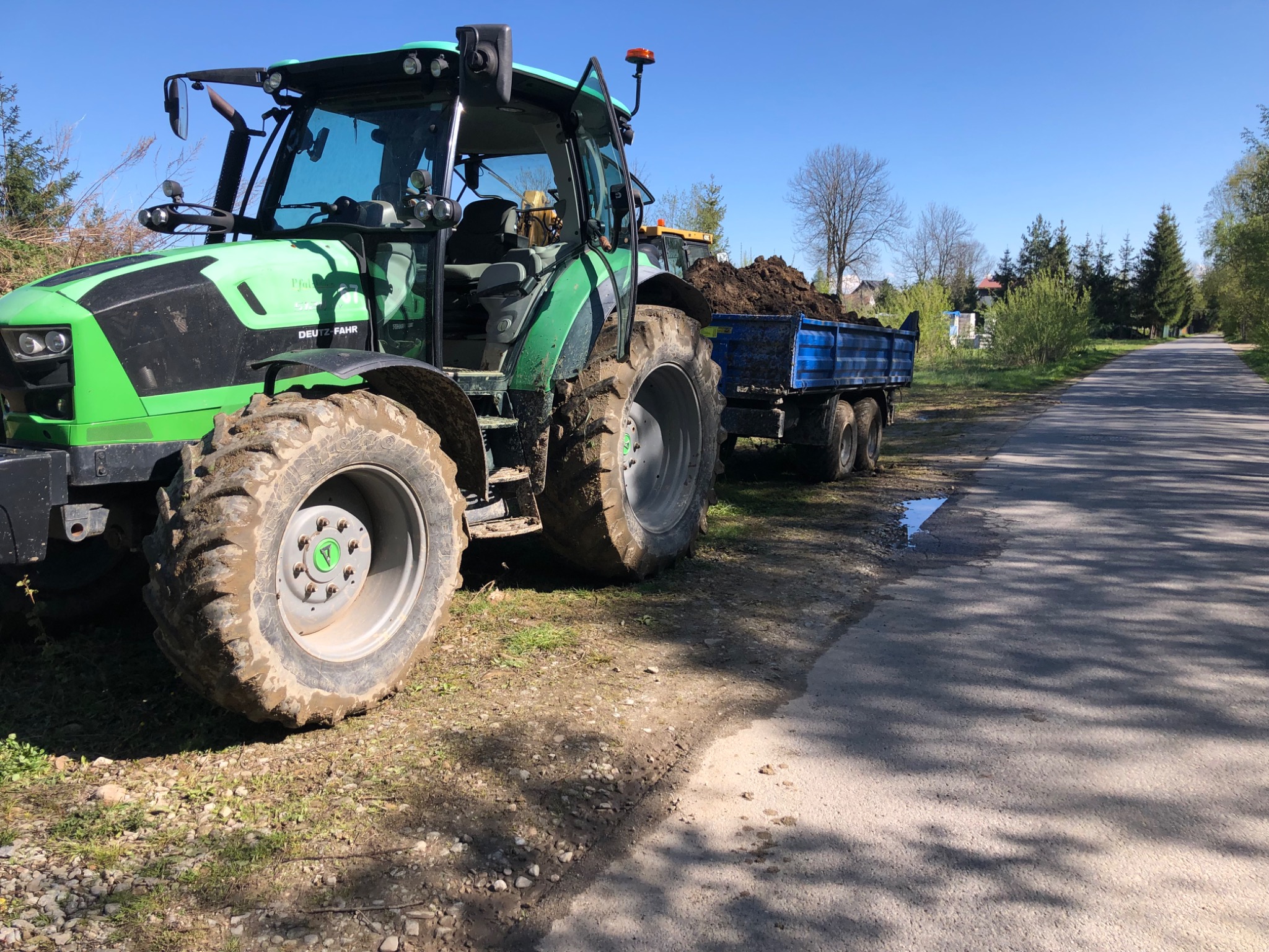 Zielony ciągnik Deutz-Fahr z przyczepą wypełnioną ziemią, stojący na poboczu asfaltowej drogi w słoneczny dzień. Widoczne błotniste opony i świeżo wykopana ziemia w przyczepie.