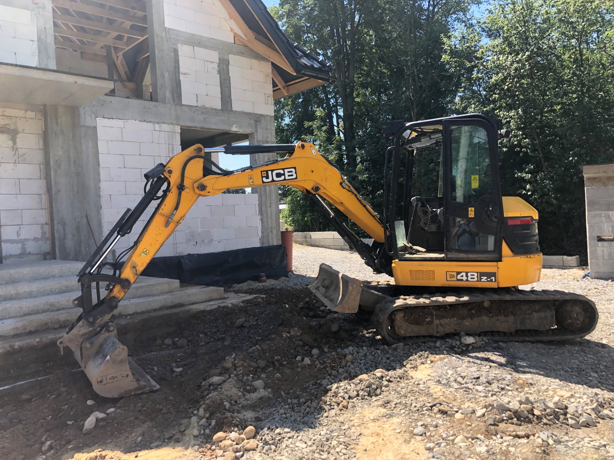 Żółta koparka JCB 48Z-1 wykonuje wykop pod fundamenty domu w stanie surowym otwartym na żwirowym podłożu, widoczne ślady pracy i świeżo wykopana ziemia.