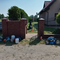 Przygotowania do prac ogrodowych: kosiarka, drabina, worki na odpady zielone i narzędzia przed ceglaną bramą wjazdową na posesję z trawnikiem i żywopłotem.