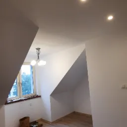 Poddasze po remoncie z panelami podłogowymi, świeżo pomalowanymi ścianami, oświetleniem sufitowym typu downlight i wiszącą lampą. Widoczne gniazdka elektryczne i pozostałości po pracach...