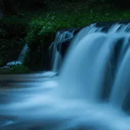 Mały wodospad w lesie, sfotografowany z długim czasem naświetlania, ukazujący rozmyte strugi wody spływające po ciemnych skałach porośniętych zielenią.