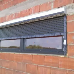 Zamknięta szara roleta zewnętrzna na oknie z dwoma szybami, zamontowana w surowej ścianie z czerwonej cegły.