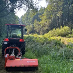 Czerwony traktor z kosiarką bijakową Agrimaster na łące, po koszeniu widoczny pokos, w tle drzewa.