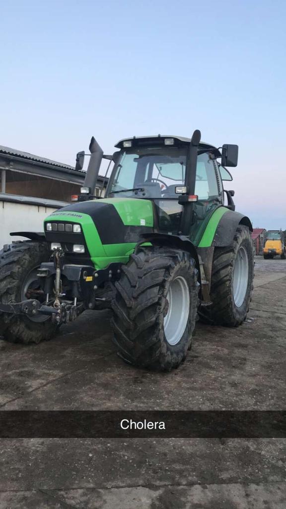 Zielono-czarny ciągnik Deutz-Fahr z dużymi oponami i szarymi felgami stoi na betonowym podłożu. W tle widoczny budynek gospodarczy i żółta ładowarka.