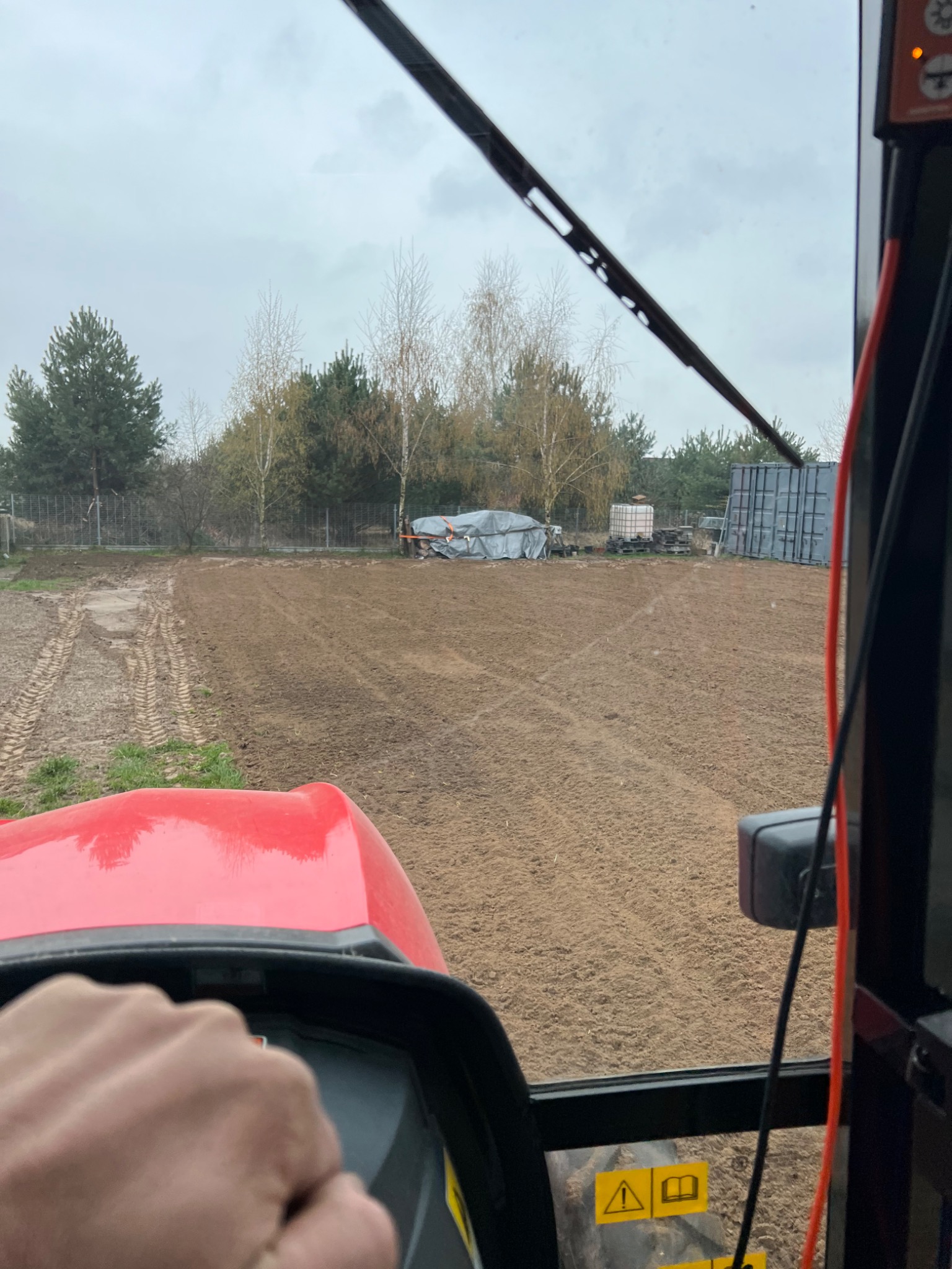 Widok z kabiny ciągnika na świeżo uprawione pole glebogryzarką separacyjną; widoczne ślady opon, w tle drzewa i zabudowania gospodarcze pod plandeką.