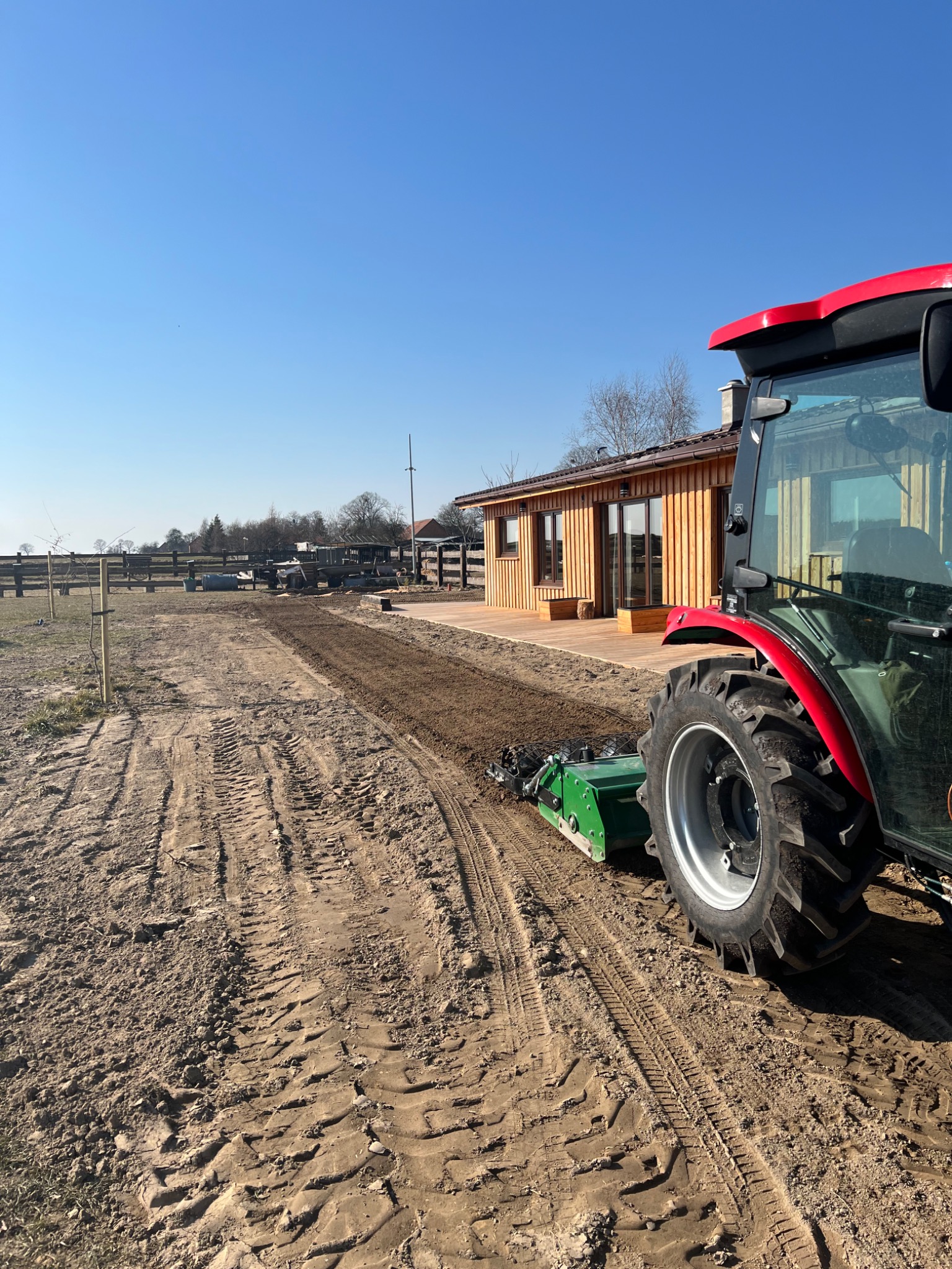 Czerwono-zielony traktor z glebogryzarką separacyjną przygotowuje ziemię pod trawnik przy drewnianym domu w słoneczny dzień. Widoczne ślady opon na piaszczystej glebie.