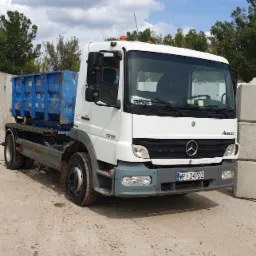 Biała ciężarówka Mercedes Atego 1218 z niebieskim kontenerem na platformie, zaparkowana na betonowym placu w słoneczny dzień. Widoczna tablica rejestracyjna WP 247D2 i naklejka 'ODPADY' za szybą.