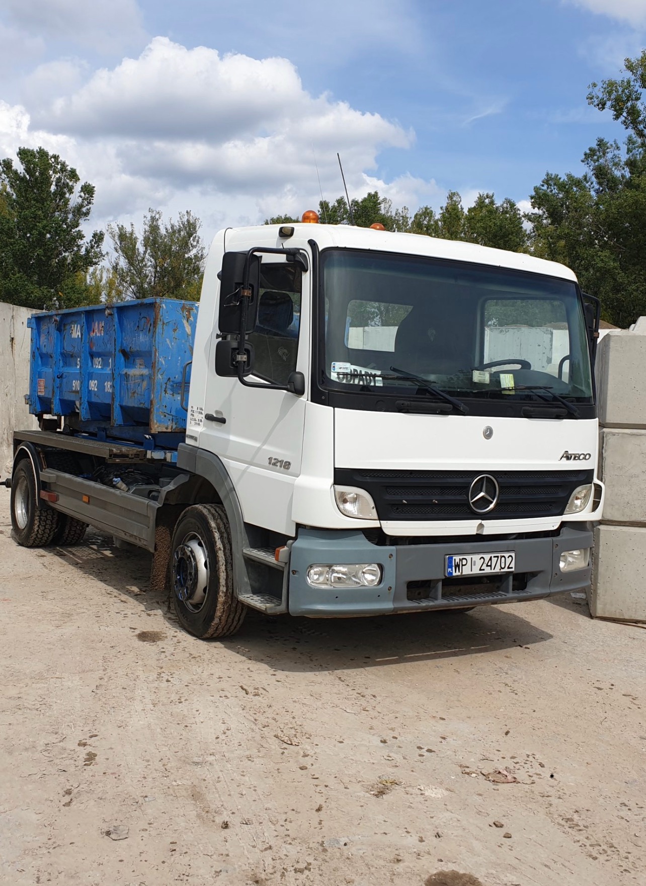 Biała ciężarówka Mercedes Atego 1218 z niebieskim kontenerem na platformie, zaparkowana na betonowym placu w słoneczny dzień. Widoczna tablica rejestracyjna WP 247D2 i naklejka 'ODPADY' za szybą.