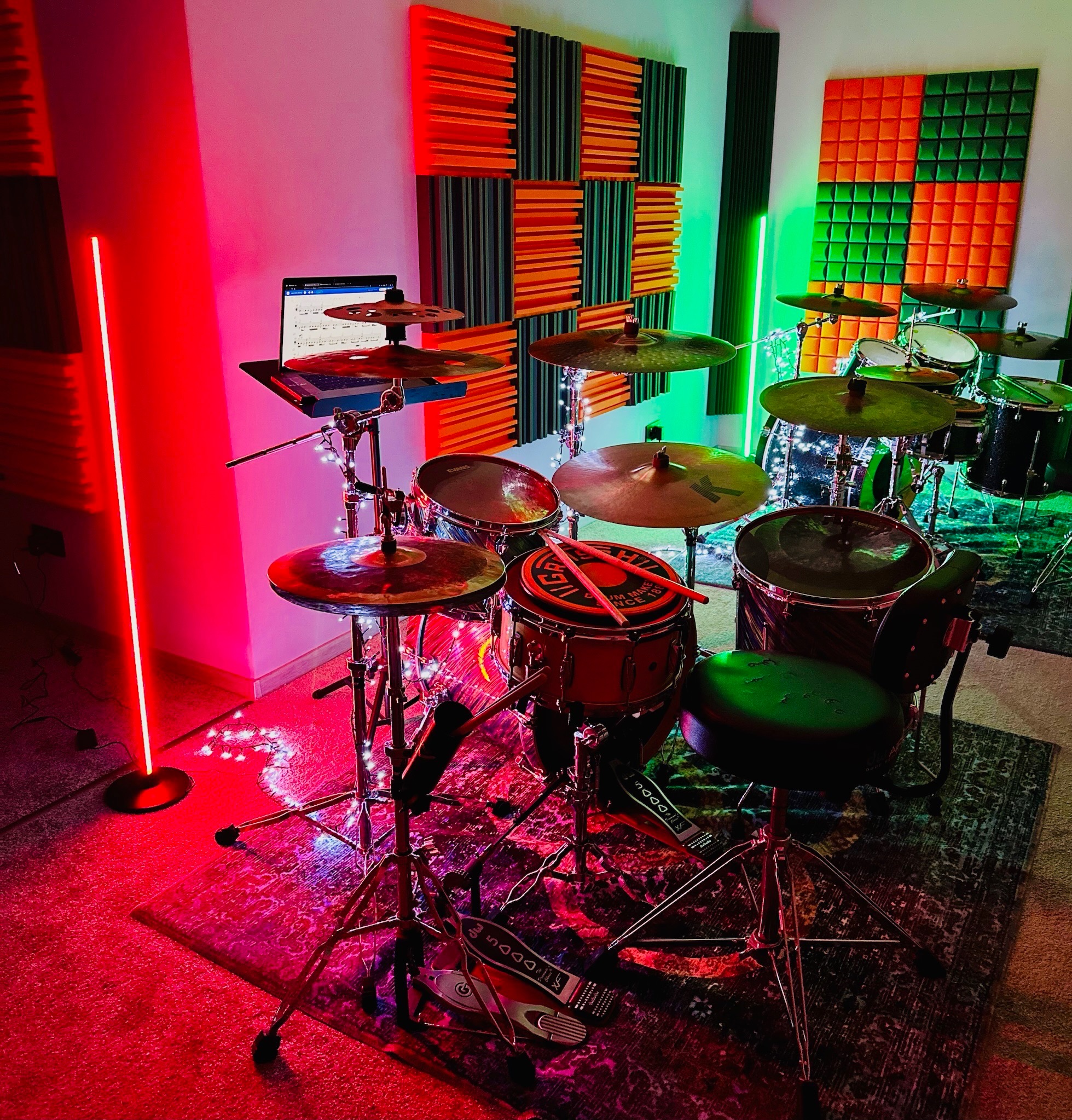 Zestaw perkusyjny w pokoju z panelami akustycznymi, oświetlony czerwonymi i zielonymi lampami LED; notatki na laptopie umieszczonym na statywie.