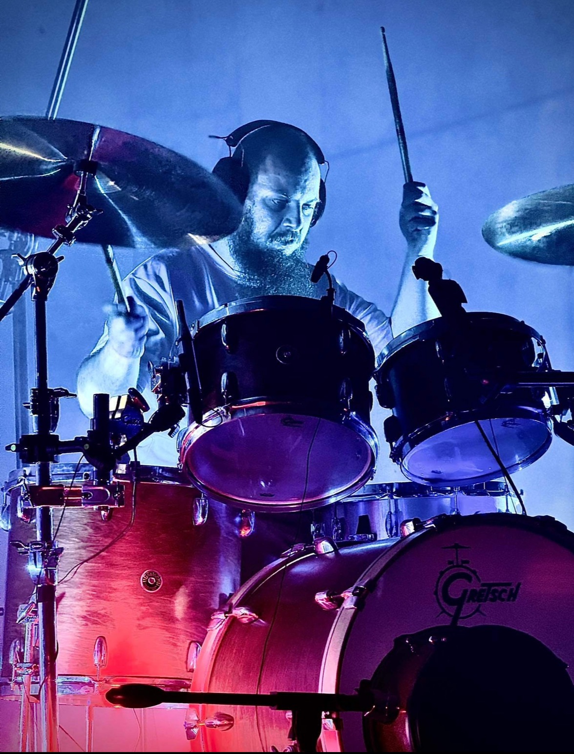 Perkusista z brodą w słuchawkach gra na perkusji Gretsch, oświetlonej niebiesko-czerwonym światłem scenicznym.