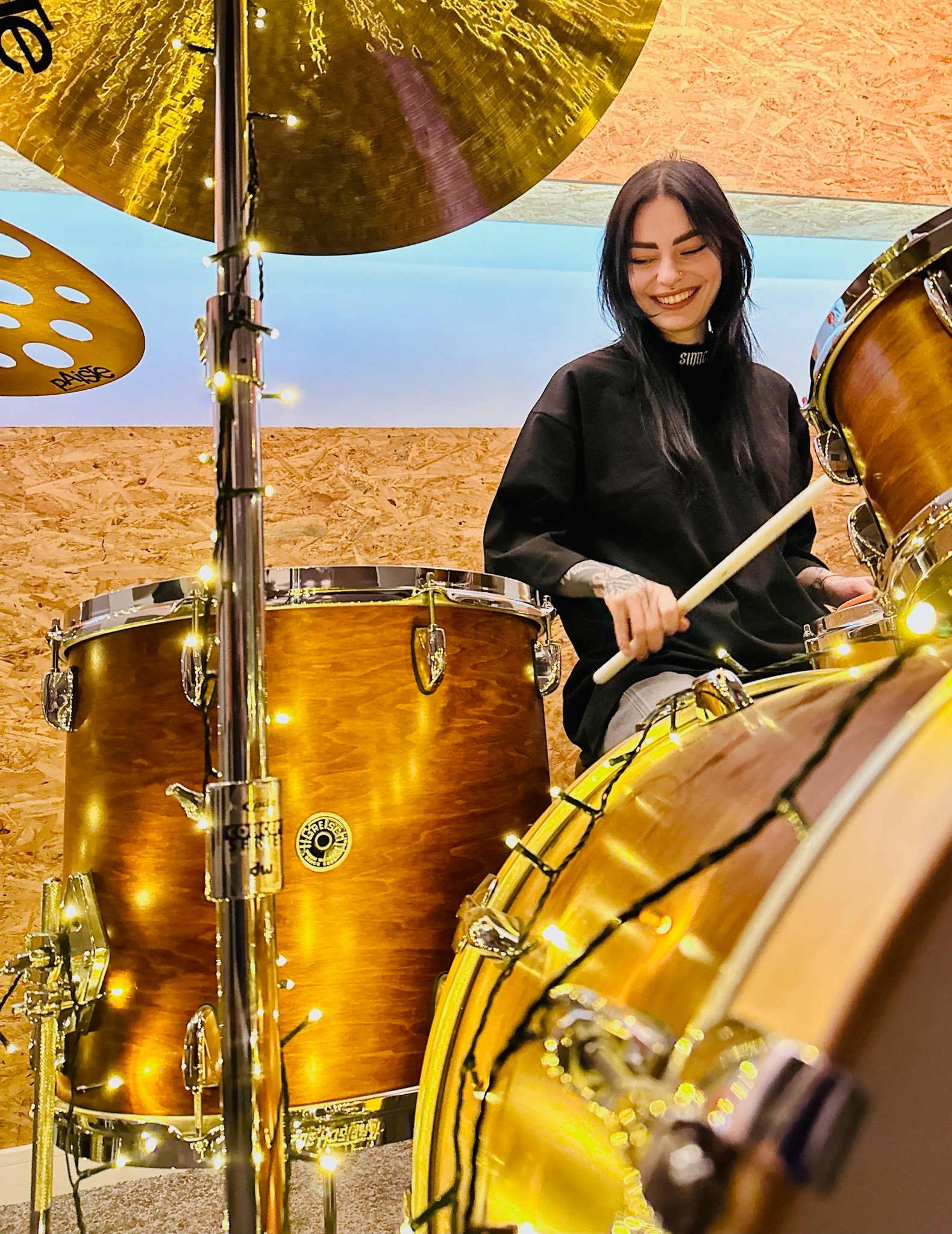 Uśmiechnięta perkusistka z tatuażami na rękach gra na perkusji Gretsch w ciepłym oświetleniu, ozdobionej światełkami.