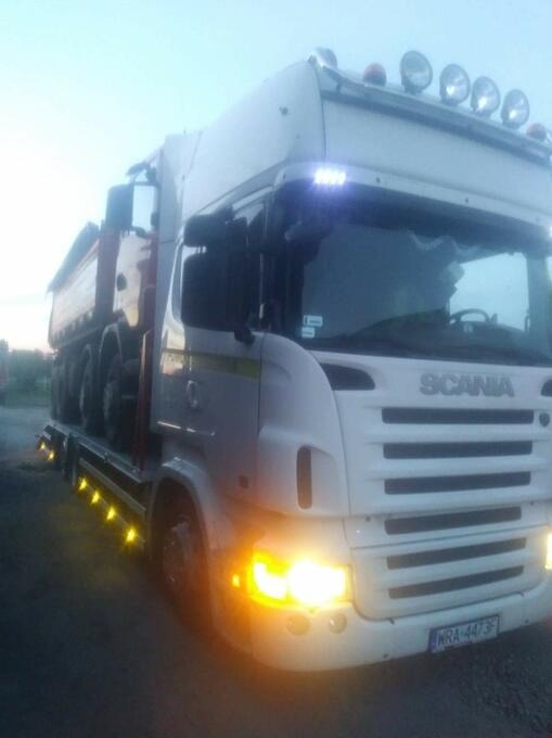 Biała ciężarówka Scania z włączonymi światłami, przewożąca inną, mniejszą ciężarówkę na platformie, widoczna tablica rejestracyjna WRA-4473F.