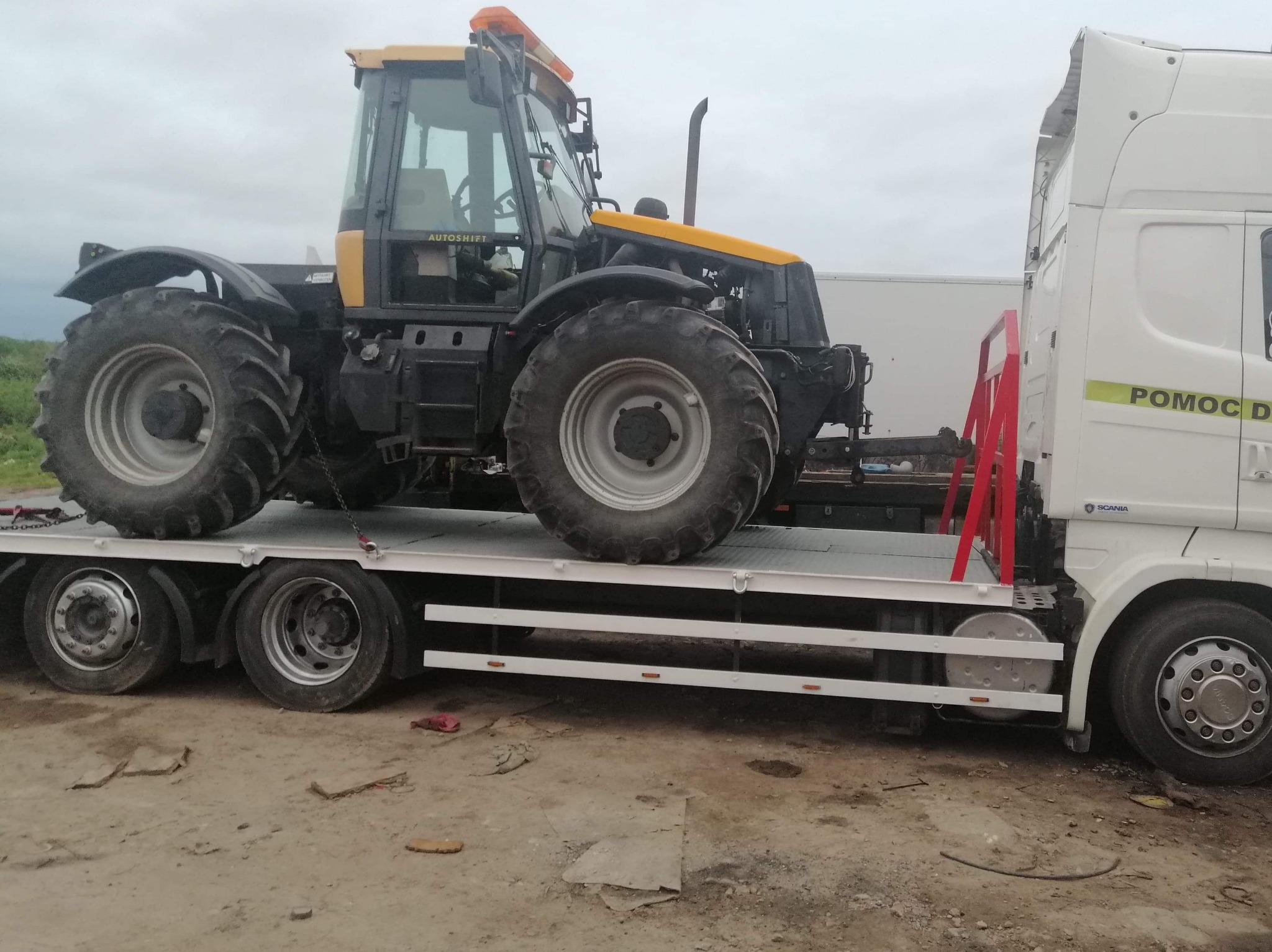 Żółto-czarny traktor rolniczy z napisem 'Autoshift' załadowany na białą lawetę z napisem 'Pomoc Drogowa' w pochmurny dzień.