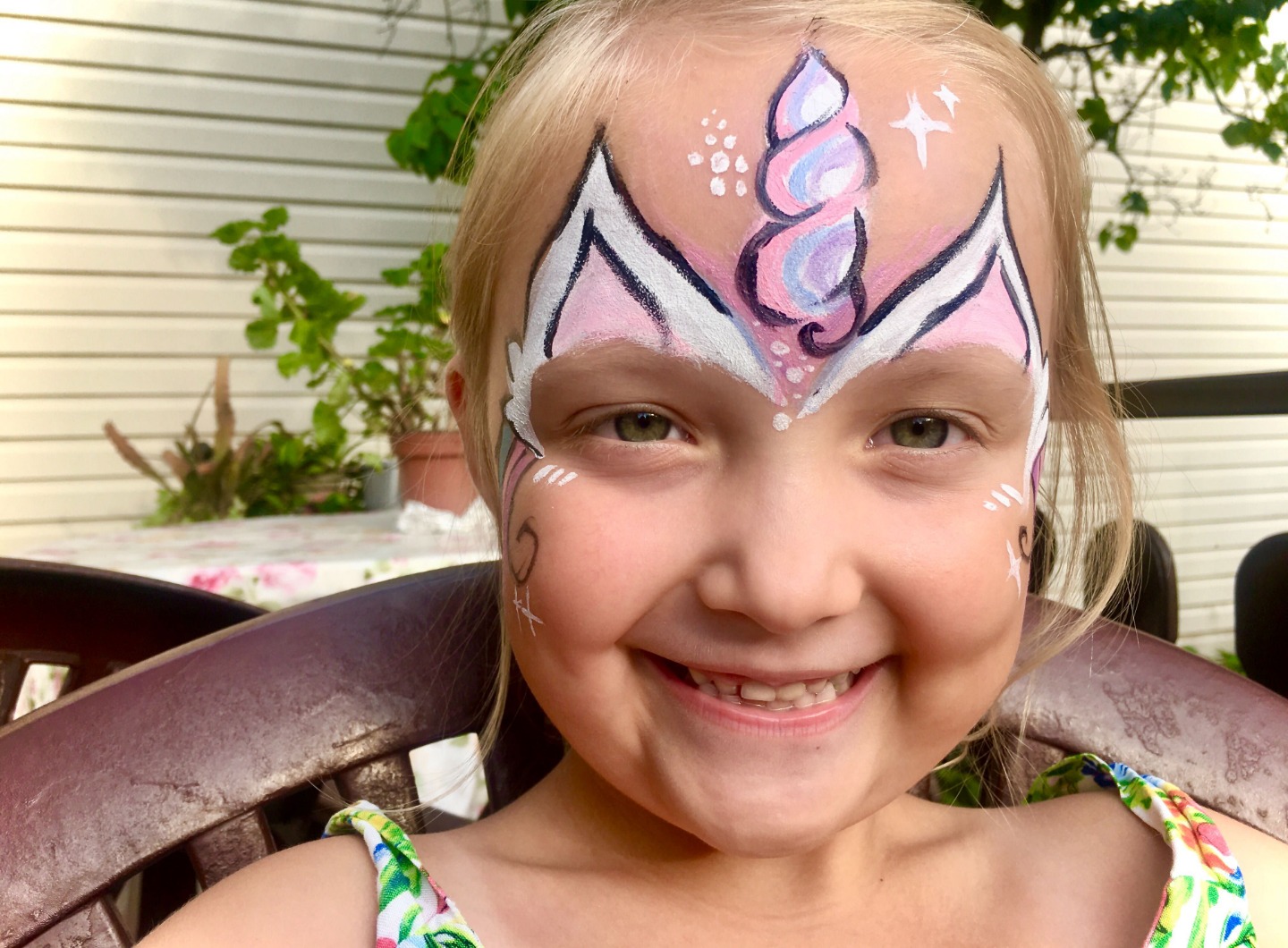 Uśmiechnięta dziewczynka z makijażem na twarzy przedstawiającym jednorożca z różowymi i fioletowymi elementami, siedząca na drewnianym krześle na tle jasnej ściany i zieleni.