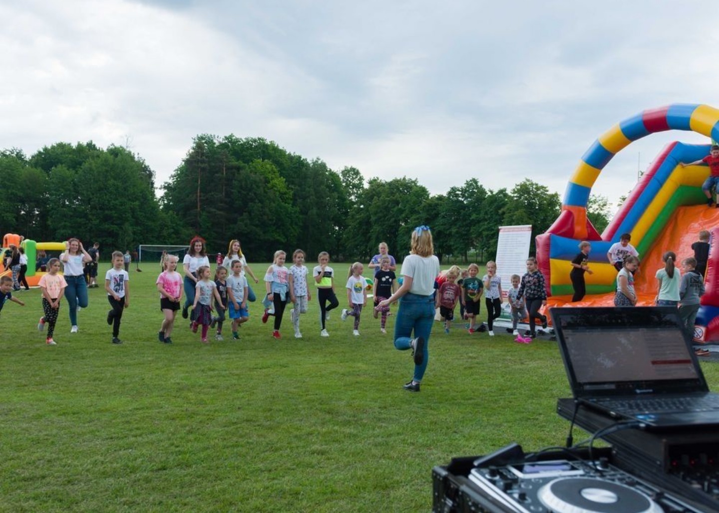 Grupa dzieci wykonuje ćwiczenia na trawie pod okiem kobiety, w tle dmuchana zjeżdżalnia i sprzęt DJ-ski.