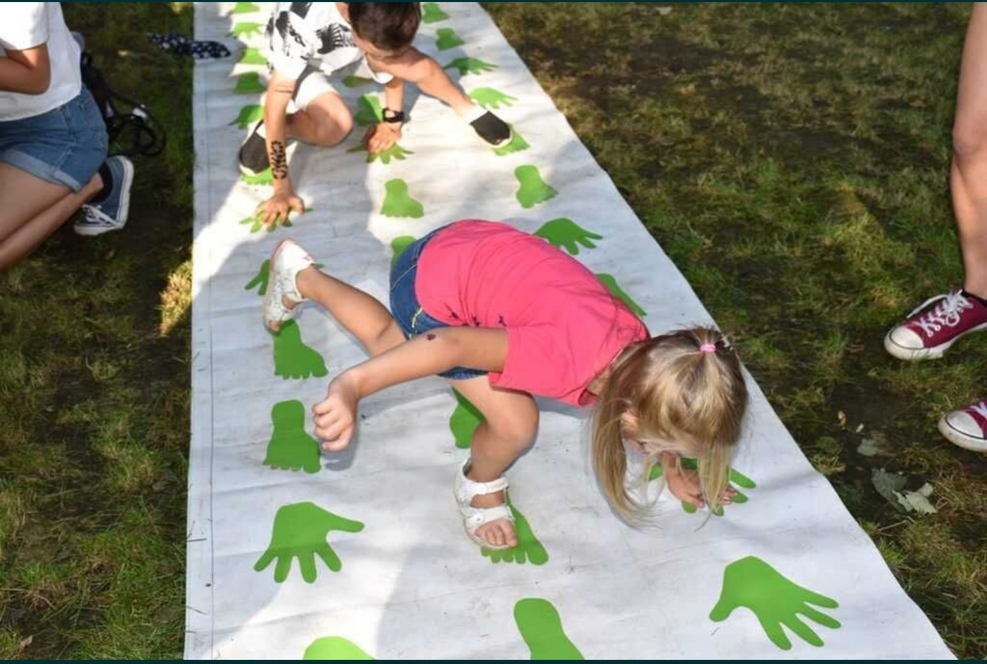 Dzieci grają w grę planszową z zielonymi odciskami dłoni i stóp na trawie, dziewczynka w różowej koszulce i sandałach próbuje utrzymać równowagę.