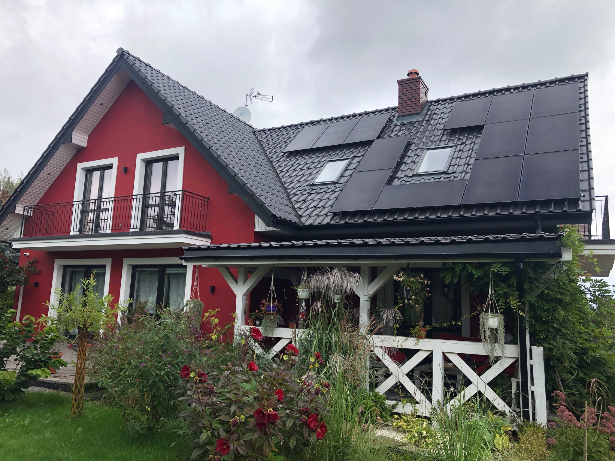 Czerwony dom jednorodzinny z ciemnoszarą dachówką, na której zamontowane są panele słoneczne; widoczna antena satelitarna i komin.