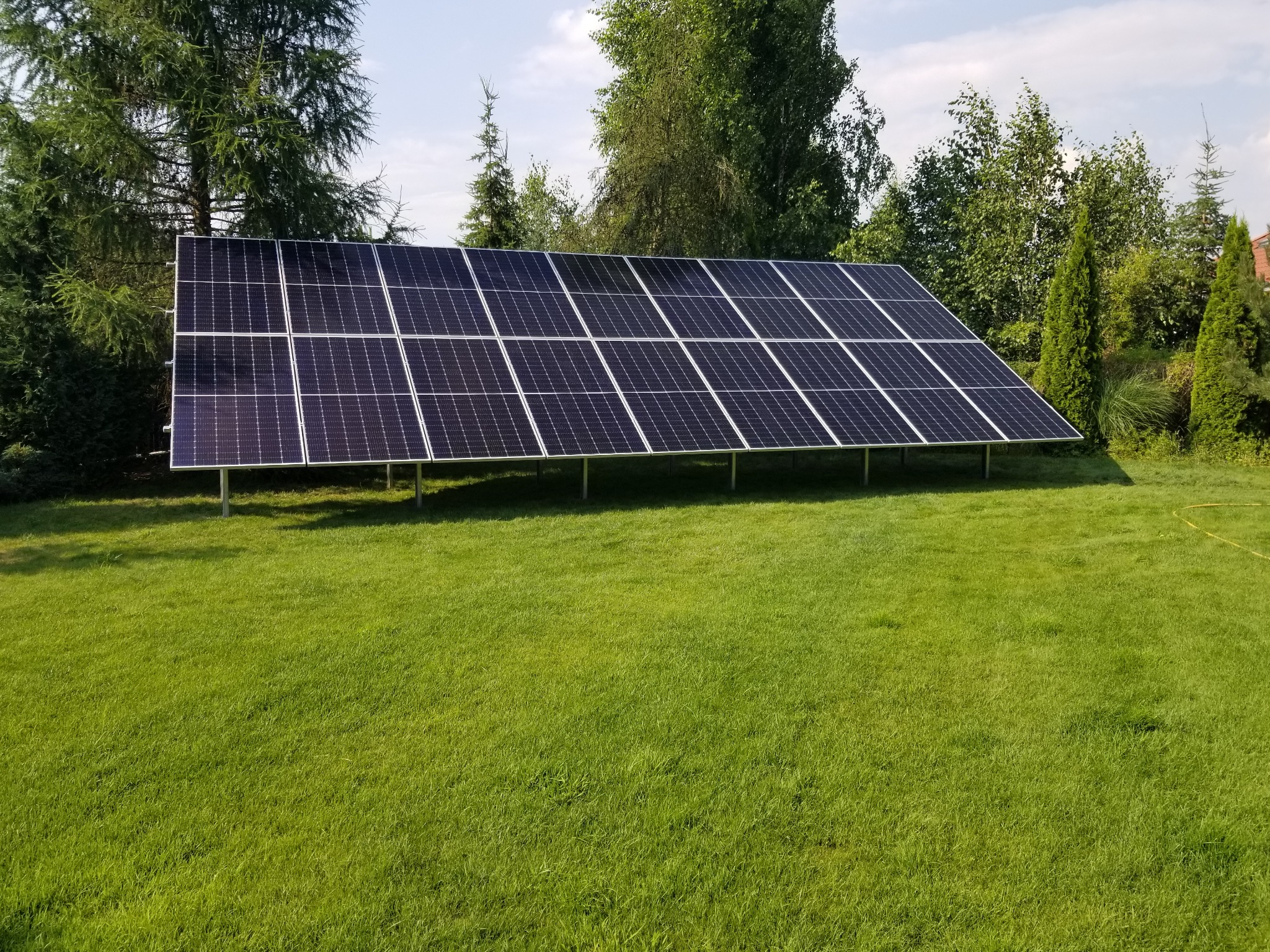 Rząd paneli słonecznych na metalowej konstrukcji, umieszczonych na trawniku przed domem, z drzewami w tle.