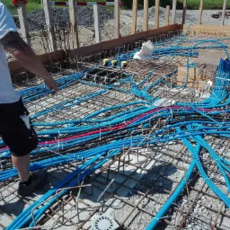 Instalacja rur karbowanych w kolorze niebieskim i czerwonym wewnątrz konstrukcji zbrojeniowej fundamentu, widoczne czarne rury z żółtymi zaślepkami i fragment sylwetki instalatora.
