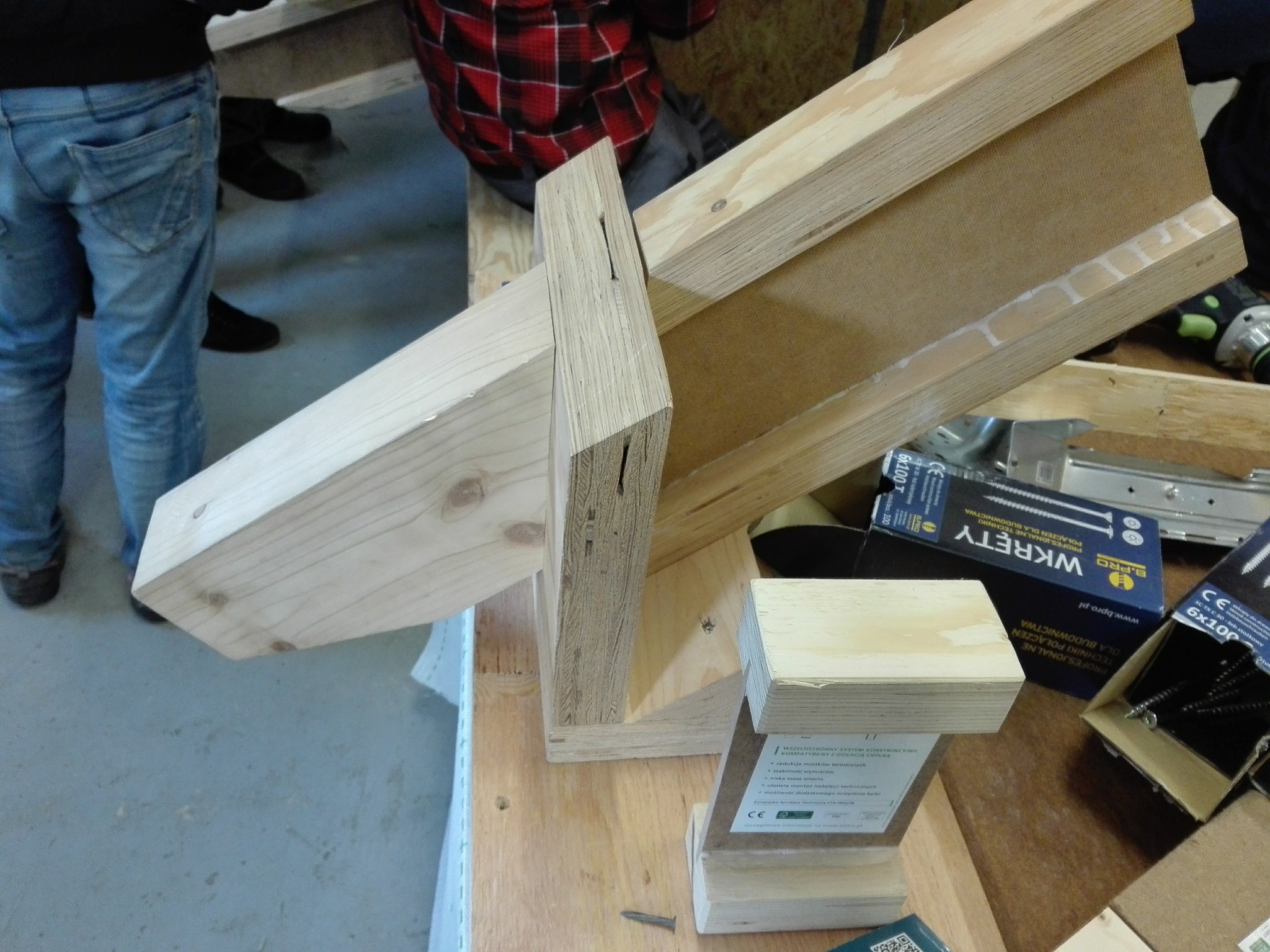 Konstrukcja z drewna i sklejki w trakcie montażu, widoczne elementy łączące oraz narzędzia, fragmenty osób w tle, warsztatowy nieład.