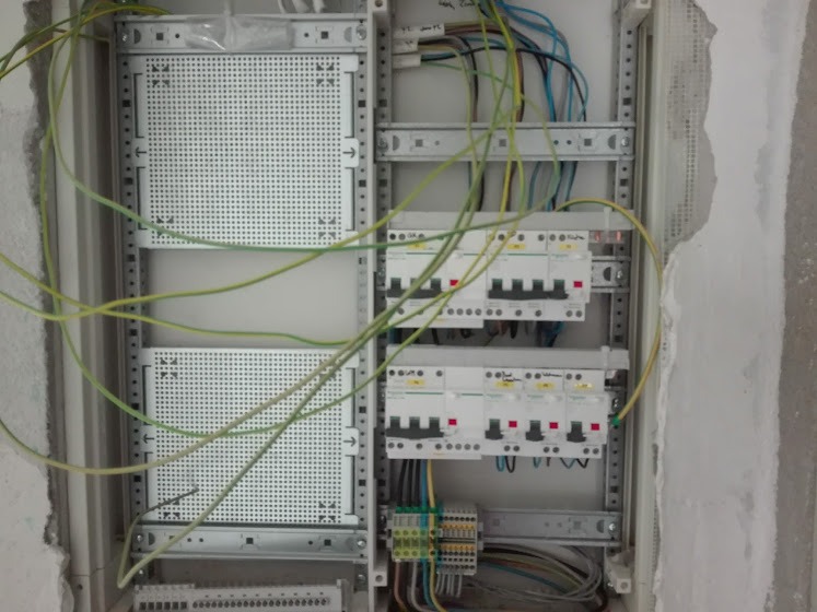 Otwarta skrzynka elektryczna w trakcie montażu, widoczne zabezpieczenia różnicowoprądowe, przewody w kolorach, metalowa perforowana płyta montażowa, surowy tynk wokół.
