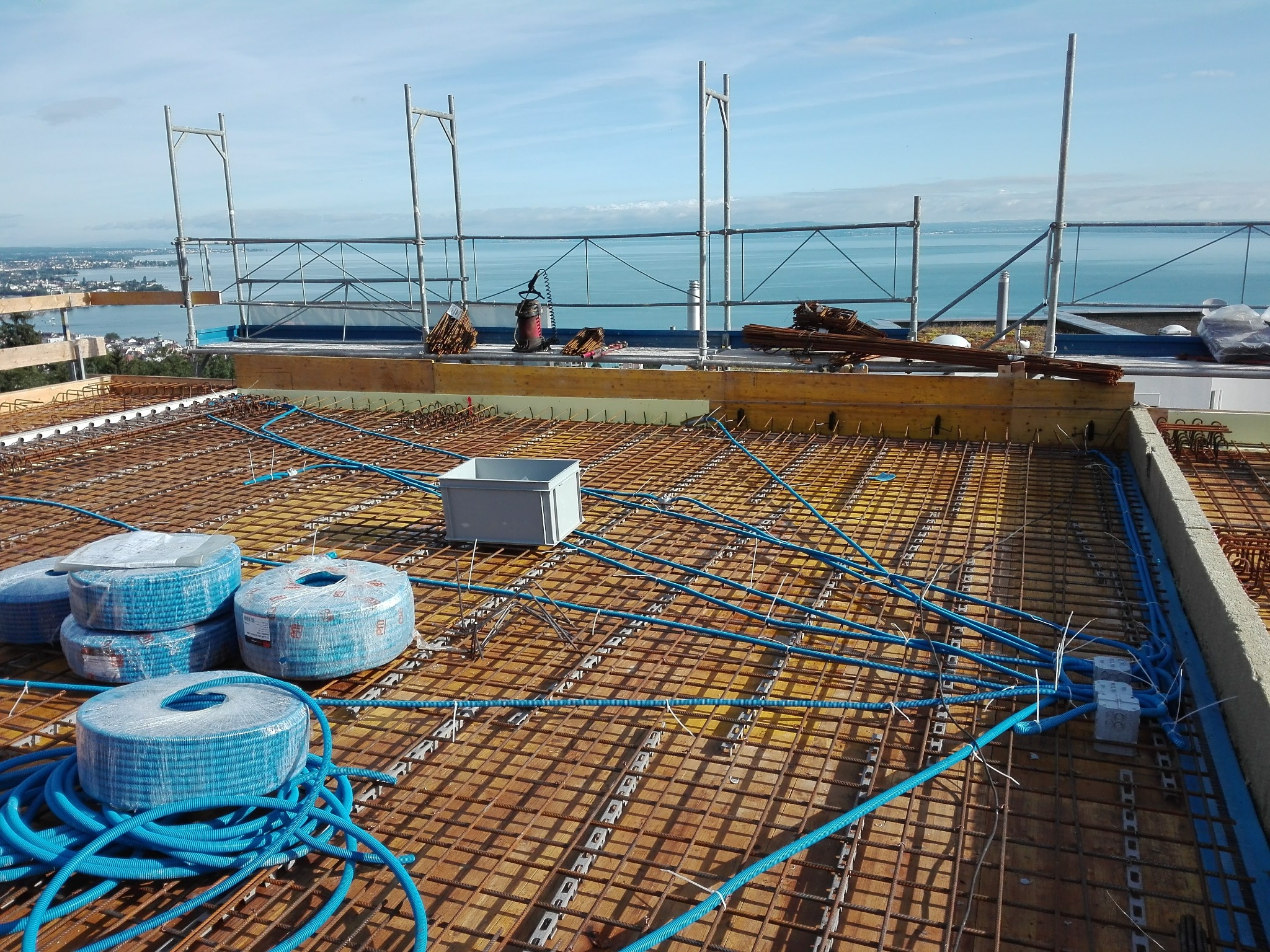 Instalacja elektryczna w rurach karbowanych, ułożona na zbrojeniu stropu, widok na jezioro i rusztowania w tle.