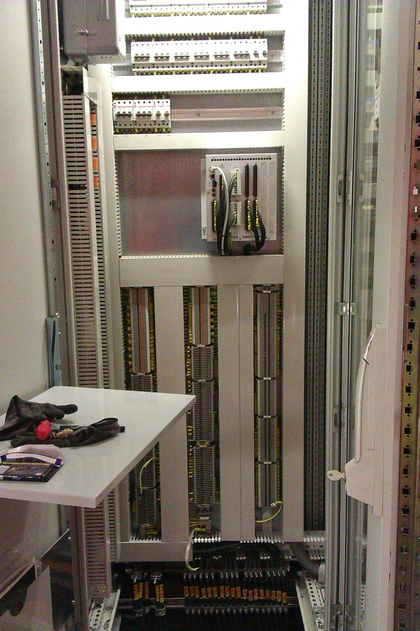 Otwarta szafa sterownicza z widocznymi elementami elektrycznymi, przewodami, listwami zaciskowymi, zabezpieczeniami, z rękawicami roboczymi i przekąską na bocznej półce.