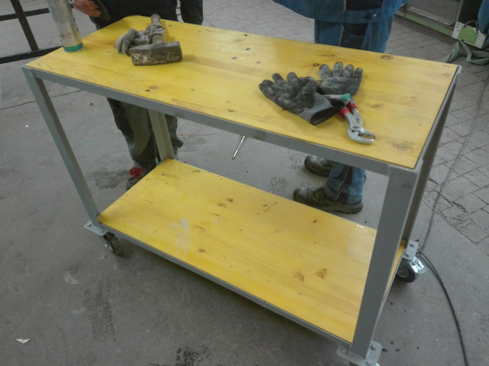 Żółty wózek warsztatowy na kółkach z metalową ramą, na blacie roboczym leżą rękawice robocze, młotek i kombinerki, obok stoi osoba w ubraniu roboczym.