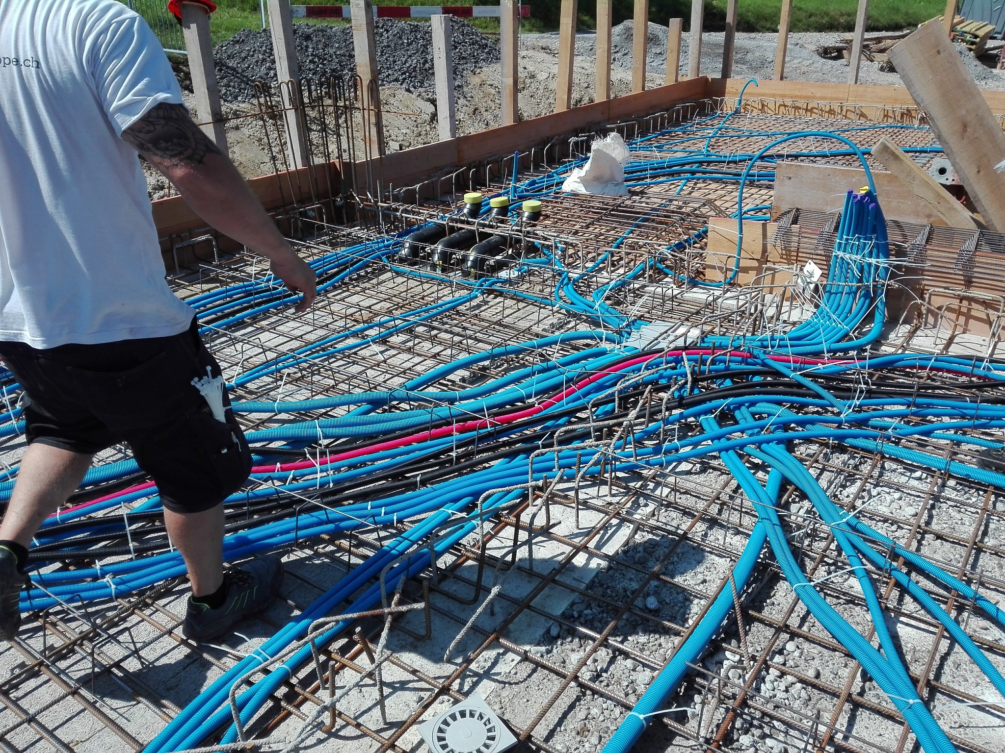 Instalacja rur karbowanych w kolorze niebieskim i czerwonym wewnątrz konstrukcji zbrojeniowej fundamentu, widoczne czarne rury z żółtymi zaślepkami i fragment sylwetki instalatora.