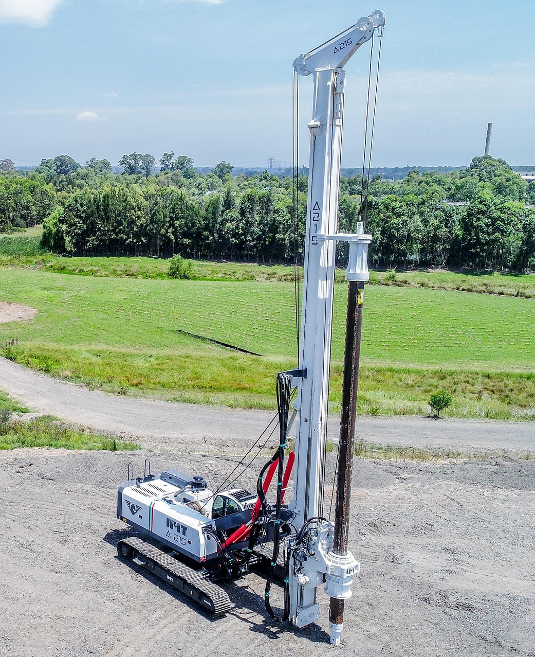 Biała wiertnica IMT A215 na gąsienicach, widok z góry na tle zieleni i błękitnego nieba. Maszyna stoi na żwirowym podłożu, gotowa do pracy.