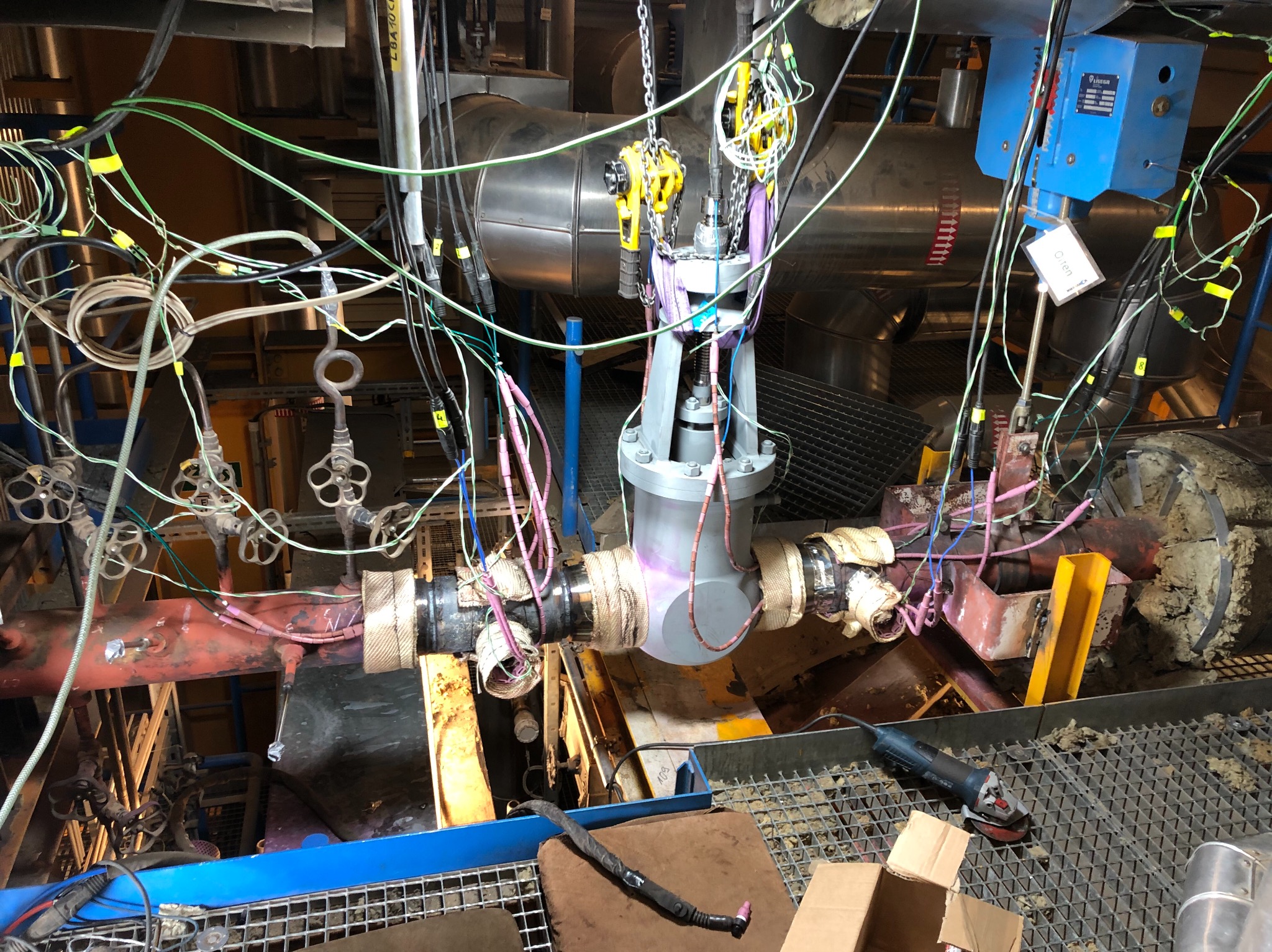 Montaż lub wymiana zaworu przemysłowego na rurociągu, widoczne zawiesie łańcuchowe, izolacja termiczna rur, liczne kable i czujniki pomiarowe oraz szlifierka kątowa na metalowej kratownicy podłoża.