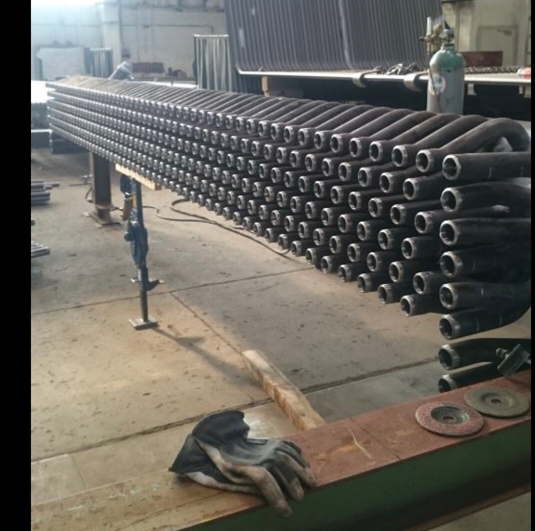 Konstrukcja z rzędów giętych rur stalowych, podparta na stojakach, widoczna w hali produkcyjnej z elementami wyposażenia, obok leżą rękawice robocze i tarcze do cięcia.