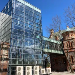 Szklana konstrukcja łącząca nowoczesny budynek z historycznym budynkiem z czerwonej cegły, widoczne klimatyzatory na zewnątrz.