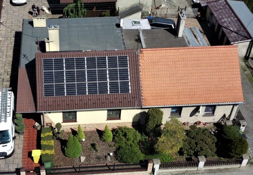 Widok z góry na dom z brązowym dachem, na którym zainstalowano panele słoneczne; obok domu zaparkowany biały samochód dostawczy z drabiną na dachu.