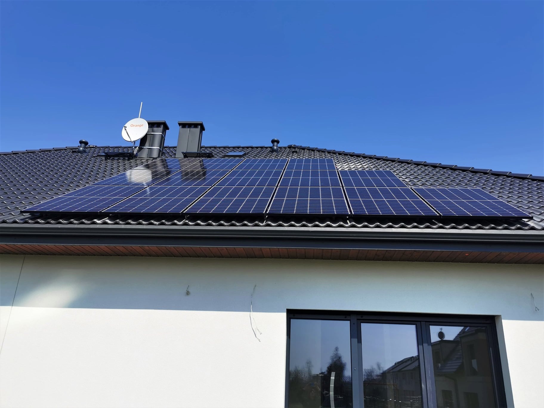 Panele słoneczne na dachu domu jednorodzinnego z anteną satelitarną, widoczne kominy i okno.