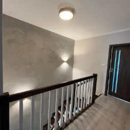 Ściana schodów pokryta dekoracyjną szarą gładzią imitującą marmur, oświetlona kinkietami, z drewnianą balustradą i widoczną częścią schodów.