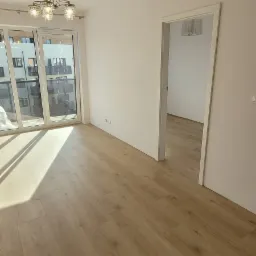 Jasne panele podłogowe ułożone w nowym mieszkaniu, widok na balkon z dużymi oknami i fragment drugiego pokoju przez otwarte drzwi.