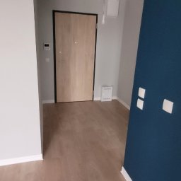 Fenix Grzegorz Woźniak - Nowe panele podłogowe w odcieniach jasnego drewna, świeżo pomalowana granatowa ściana z białymi listwami przypodłogowymi, widoczne gniazdka elektryczne i drzwi wejściowe w tle.