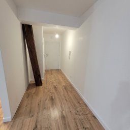 Fenix Grzegorz Woźniak - Wnętrze mieszkania po remoncie: drewnopodobne panele podłogowe ułożone wzdłuż korytarza z białymi ścianami i widoczną drewnianą belką konstrukcyjną.