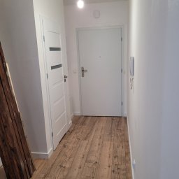 Fenix Grzegorz Woźniak - Korytarz po remoncie z ułożonymi panelami podłogowymi imitującymi drewno, widoczne białe drzwi wejściowe i wewnętrzne, oraz sterta drewnianych desek oparta o ścianę.