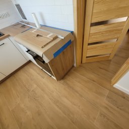 Fenix Grzegorz Woźniak - Podłoga wyłożona panelami imitującymi drewno w mieszkaniu we Wrocławiu, widoczny fragment zabudowy kuchennej z zabezpieczonym blatem i nowa drewniana ościeżnica drzwiowa.