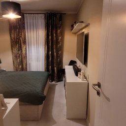 Fenix Grzegorz Woźniak - Sypialnia z łóżkiem z zieloną narzutą, zasłonami w tropikalne wzory, białymi meblami i telewizorem na ścianie, oświetlona lampą wiszącą z czarnym abażurem.