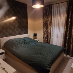 Fenix Grzegorz Woźniak - Nowoczesna sypialnia z łóżkiem kontynentalnym, ciemnozieloną narzutą i lampą sufitową z abażurem, widoczne tapety w motywy roślinne i zasłony w tropikalne wzory.