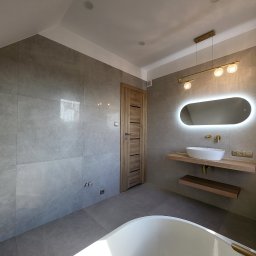 Fenix Grzegorz Woźniak - Nowoczesna łazienka z szarymi płytkami, podświetlanym owalnym lustrem nad umywalką na drewnianym blacie, złotą armaturą i lampą, oraz fragmentem białej wanny.