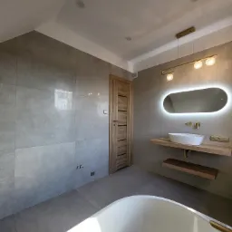 Nowoczesna łazienka z szarymi płytkami, podświetlanym owalnym lustrem nad umywalką na drewnianym blacie, złotą armaturą i lampą, oraz fragmentem białej wanny.