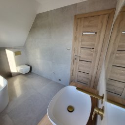 Fenix Grzegorz Woźniak - Nowoczesna łazienka z umywalką nablatową, baterią w kolorze złotym, szarą glazurą, drewnianymi drzwiami i wiszącą toaletą.