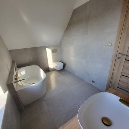 Fenix Grzegorz Woźniak - Nowoczesna łazienka na poddaszu z wanną wolnostojącą, umywalką nablatową z złotymi akcentami i szarą płytką wielkoformatową na ścianach.